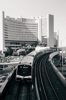 Black and white photo of a train on elevated tracks passing modern buildings in a cityscape.