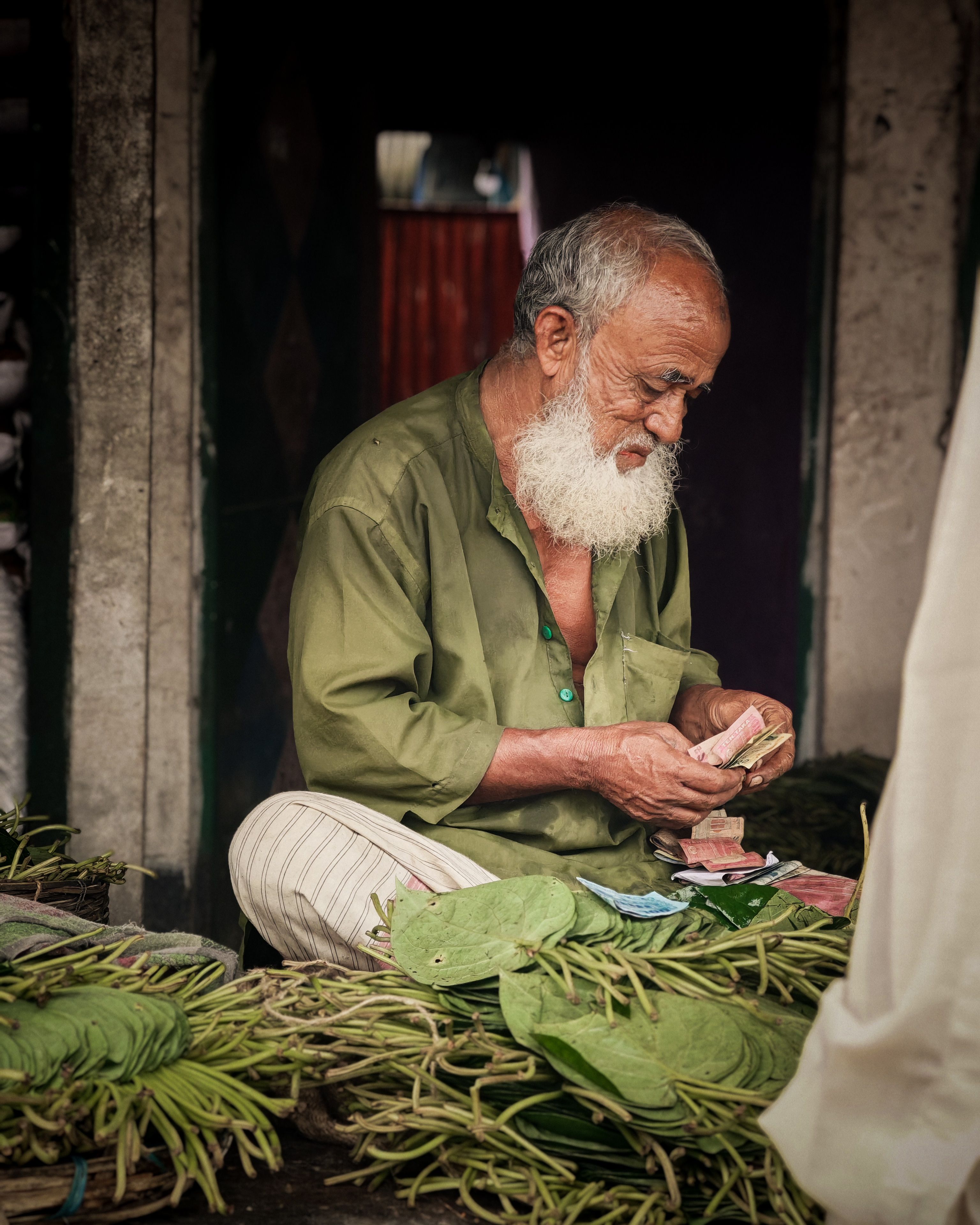 An elderly vendor counts money at an outdoor market, surrounded by green betel leaves.