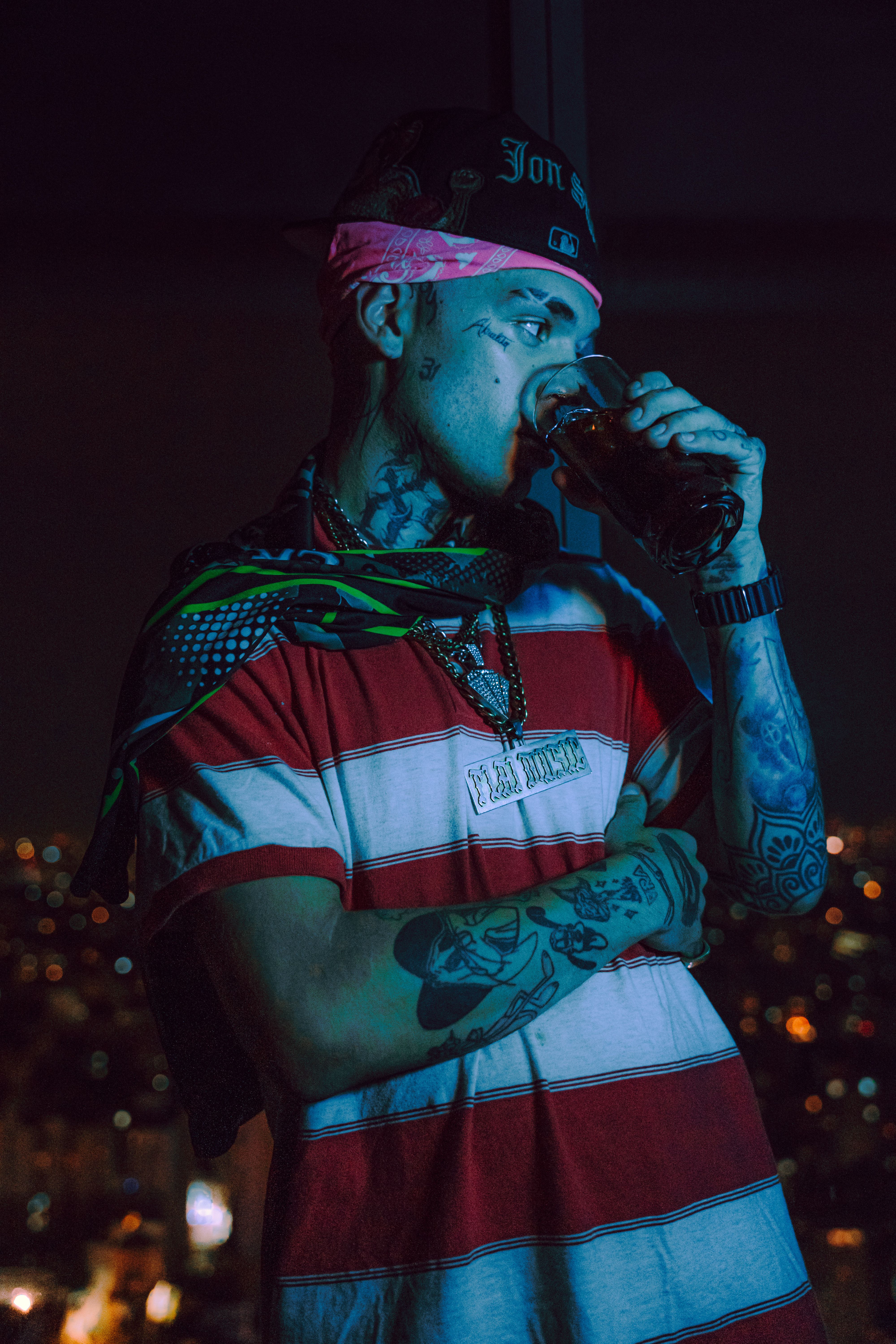 A tattooed artist in a striped shirt enjoys a drink at night, overlooking city lights.