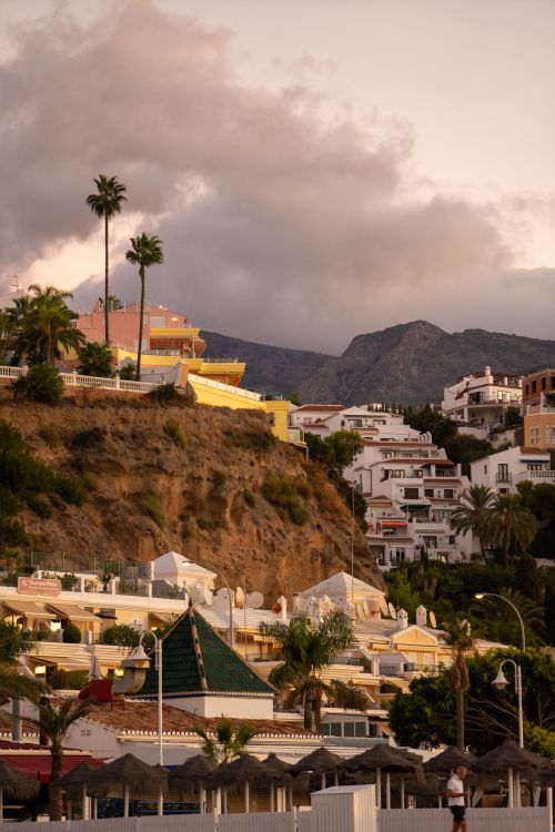 Free Charming hillside houses in Nerja, Spain with a mountain backdrop during sunset. Stock Photo