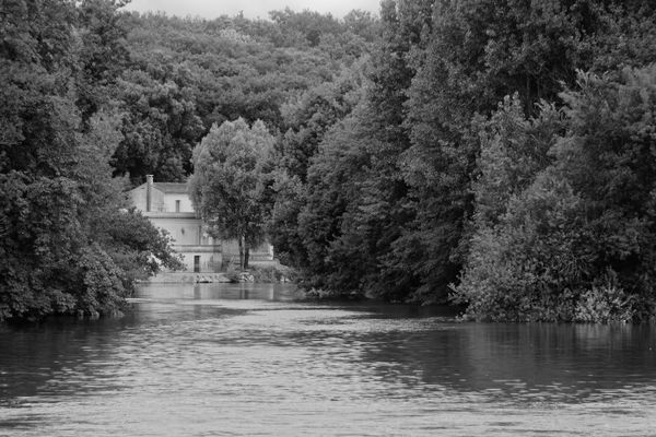 Black and white image of a secluded house by a river surrounded by lush forest