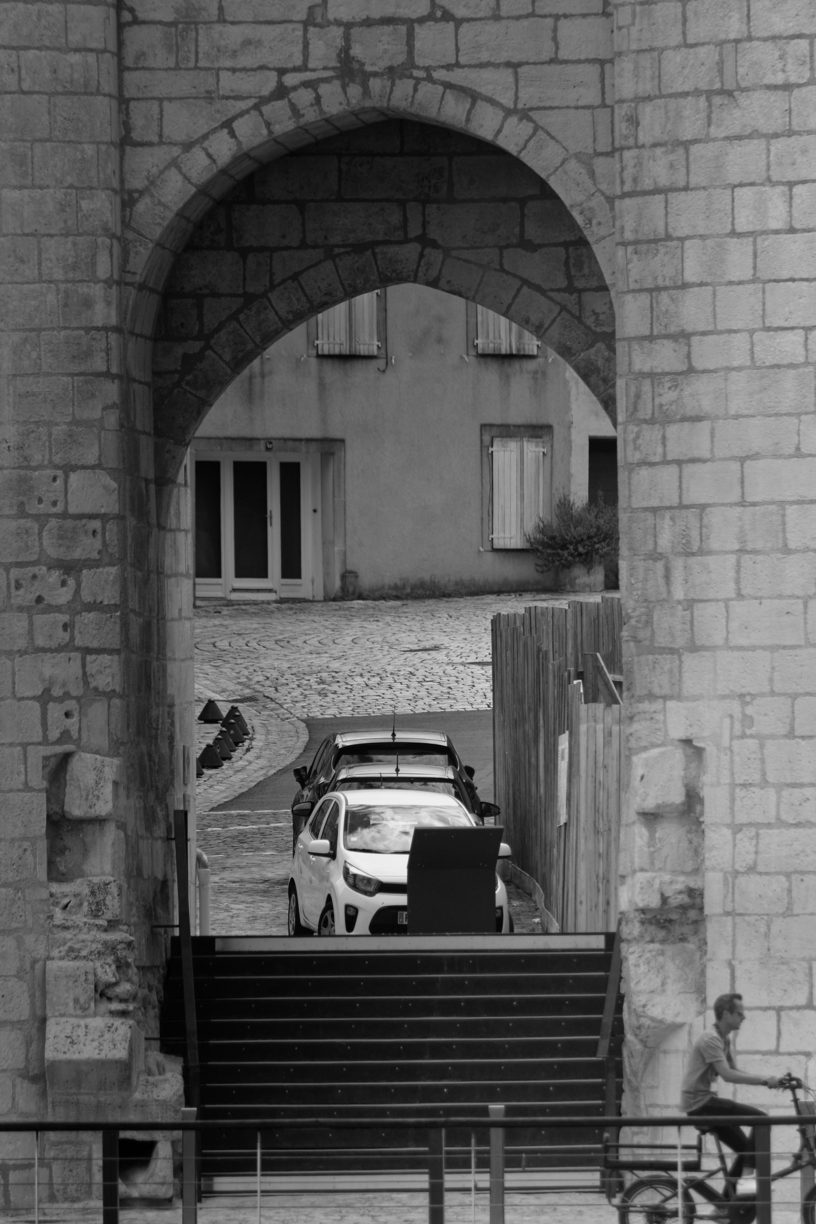 Black and white view through a historic archway showing a car and a cyclist in an urban setting