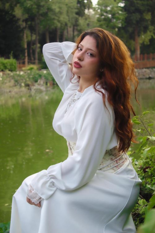 Free A woman in a white dress poses gracefully by a serene green pond. Stock Photo