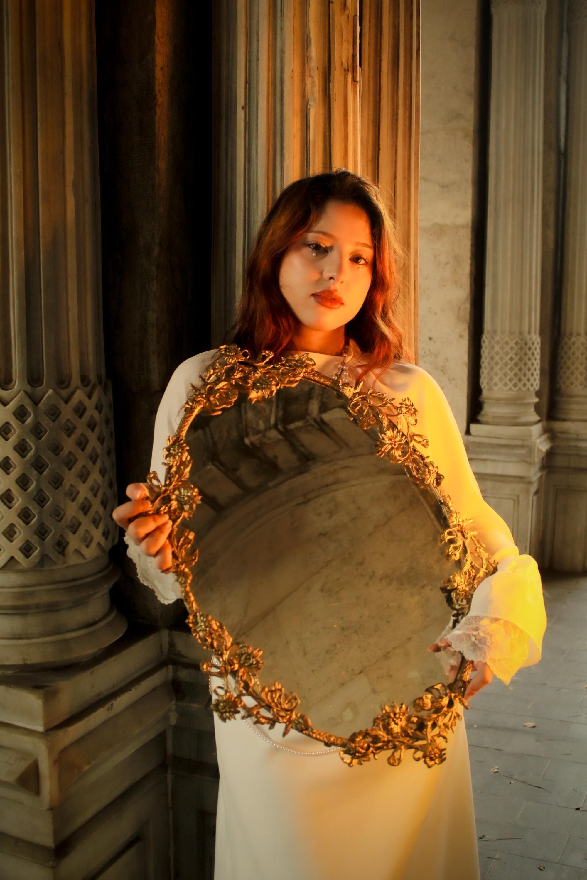 A woman in a white dress holds a vintage mirror in an elegant indoor setting.