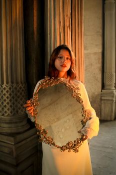 A woman in an elegant setting holds an ornate antique mirror, framed by classical architecture.