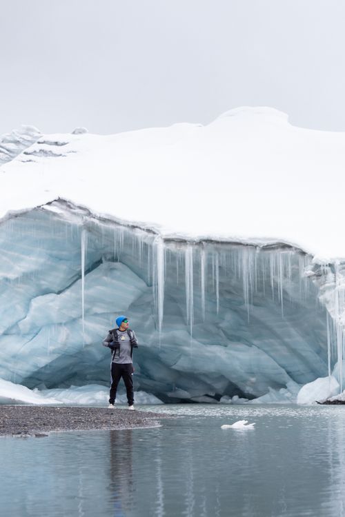 Free A person stands near a massive ice cavern with icicles hanging above a partially frozen lake. Stock Photo