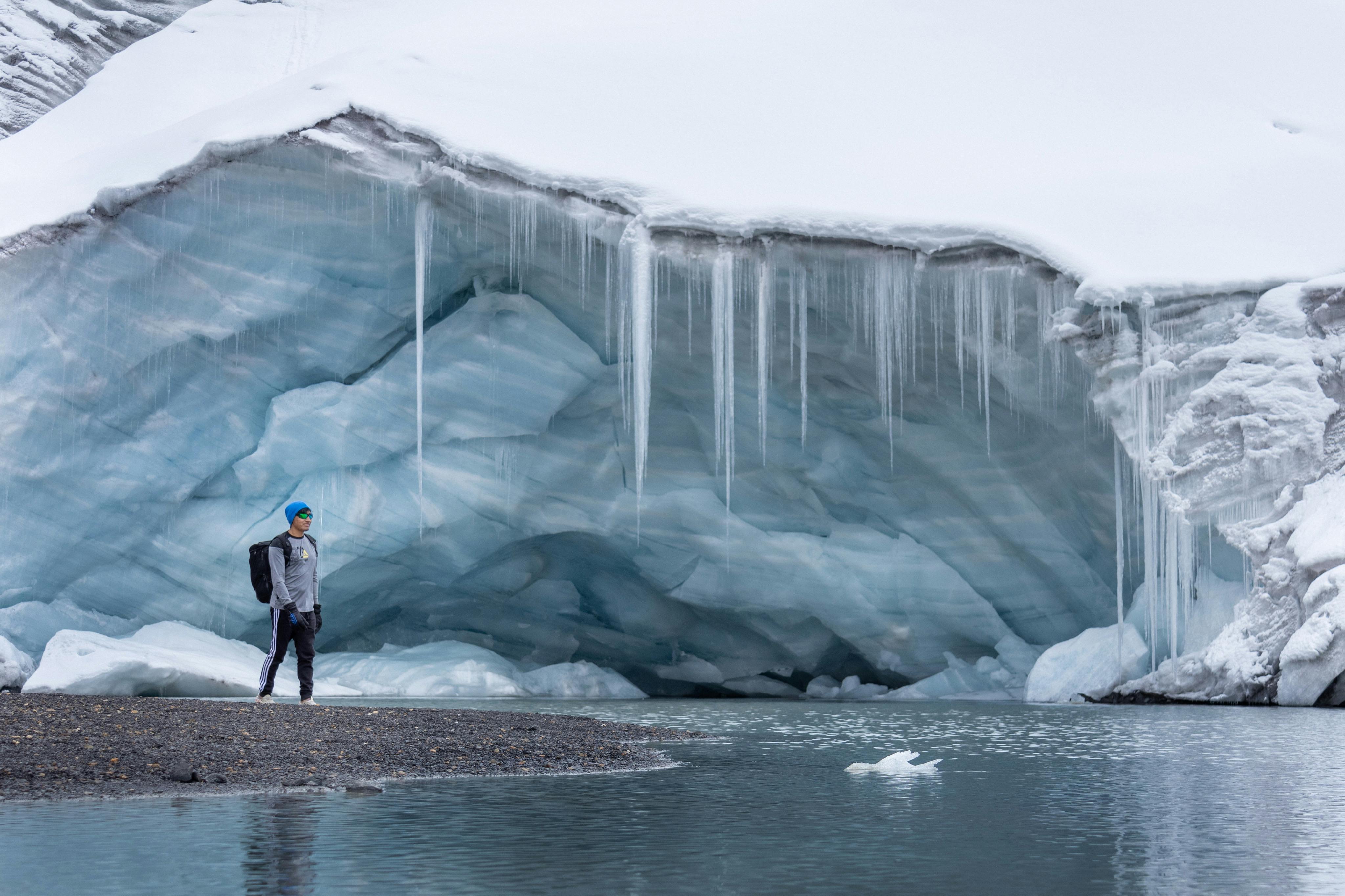 A lone explorer stands near an ice cave by a frozen lake in the Arctic, showcasing adventure.