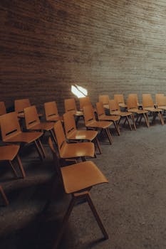 An arrangement of empty wooden chairs in a textured minimalist room with natural light.