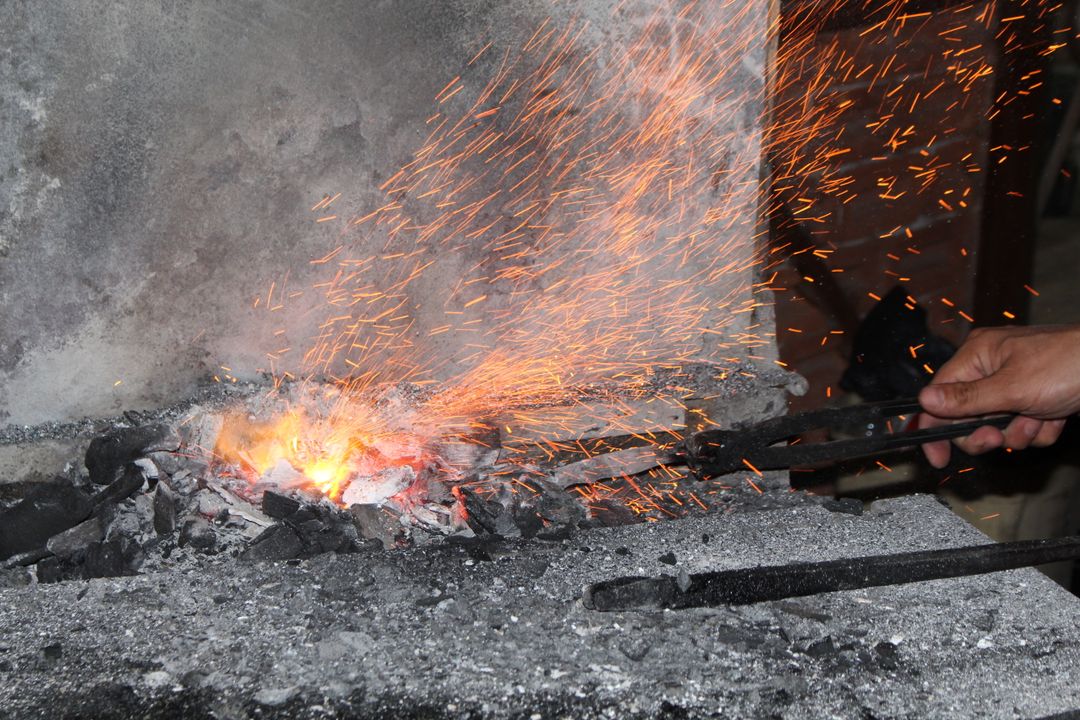 A blacksmith's hand holding long steel tongs over glowing forge coals, a dense shower of orange sparks rising against a stone wall
