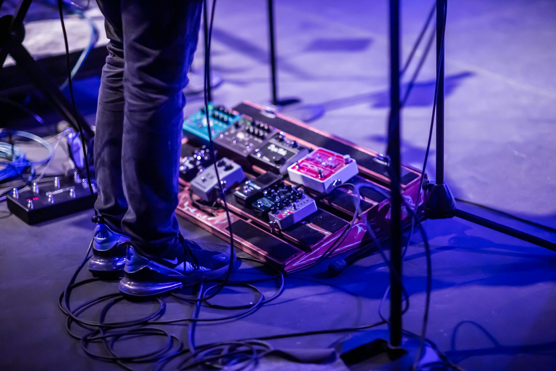 A musician performing with a pedalboard on a stage lit by blue lights. Vivid and dynamic.