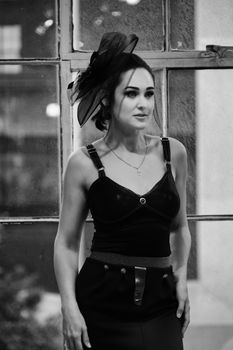 Chic woman in a stylish black outfit and hat stands by a vintage window indoors.
