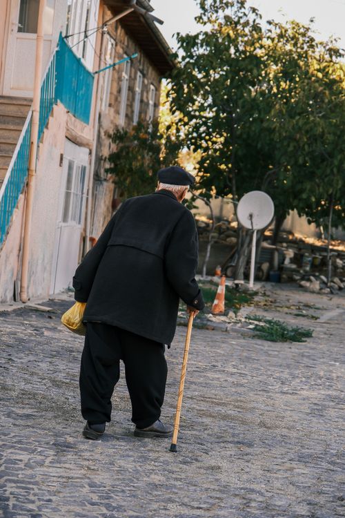 Free Elderly man in black coat walking with cane on quiet cobblestone street on sunny day. Stock Photo