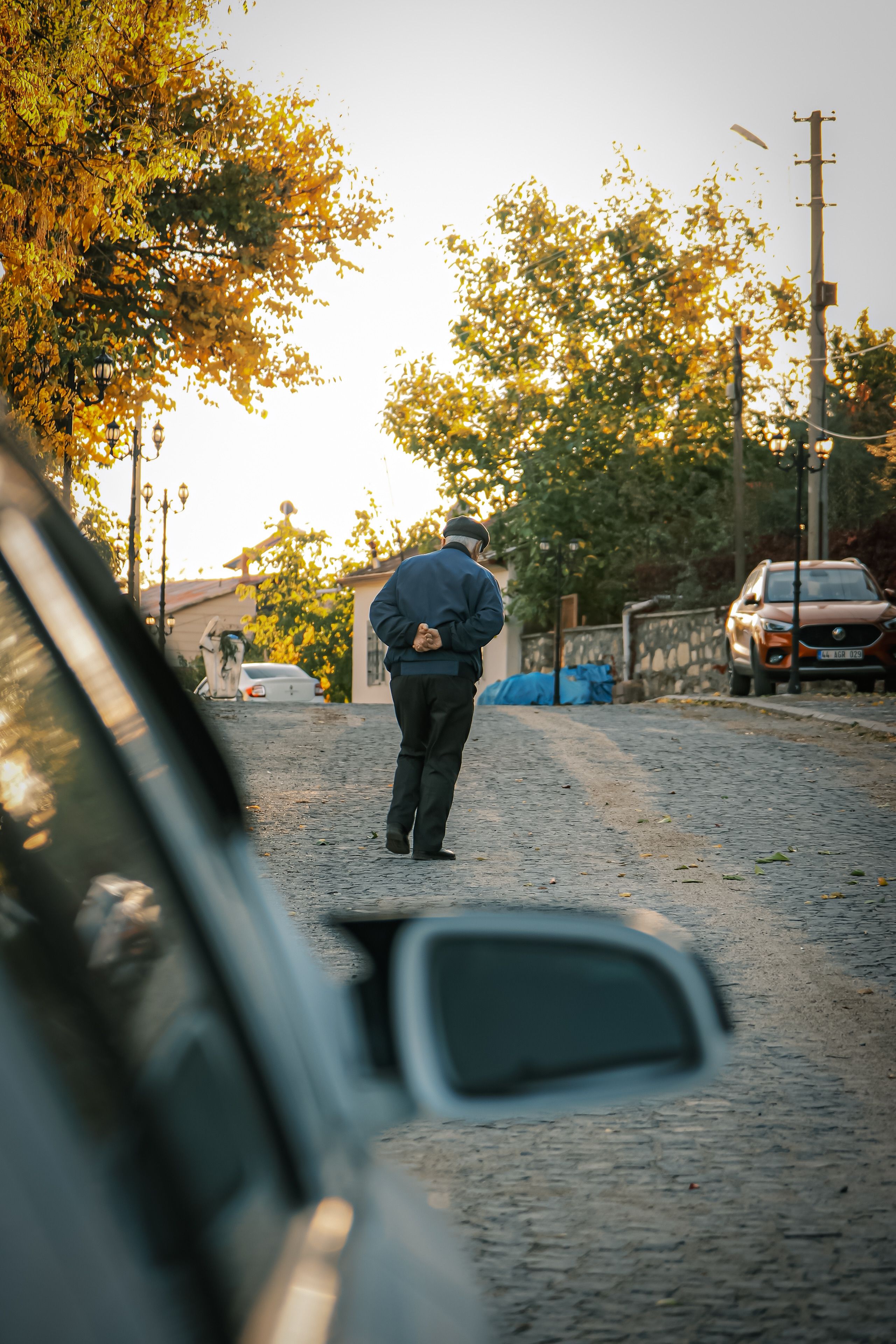 Elderly man walking down a cobblestone street during a serene sunset, with cars parked nearby.