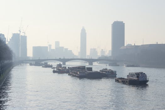 薄雾笼罩的天空下，朦胧的城市天际线旁，一条平静的河上有小船