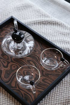 Minimalist glass teapot set on textured wooden tray with cozy rug backdrop.