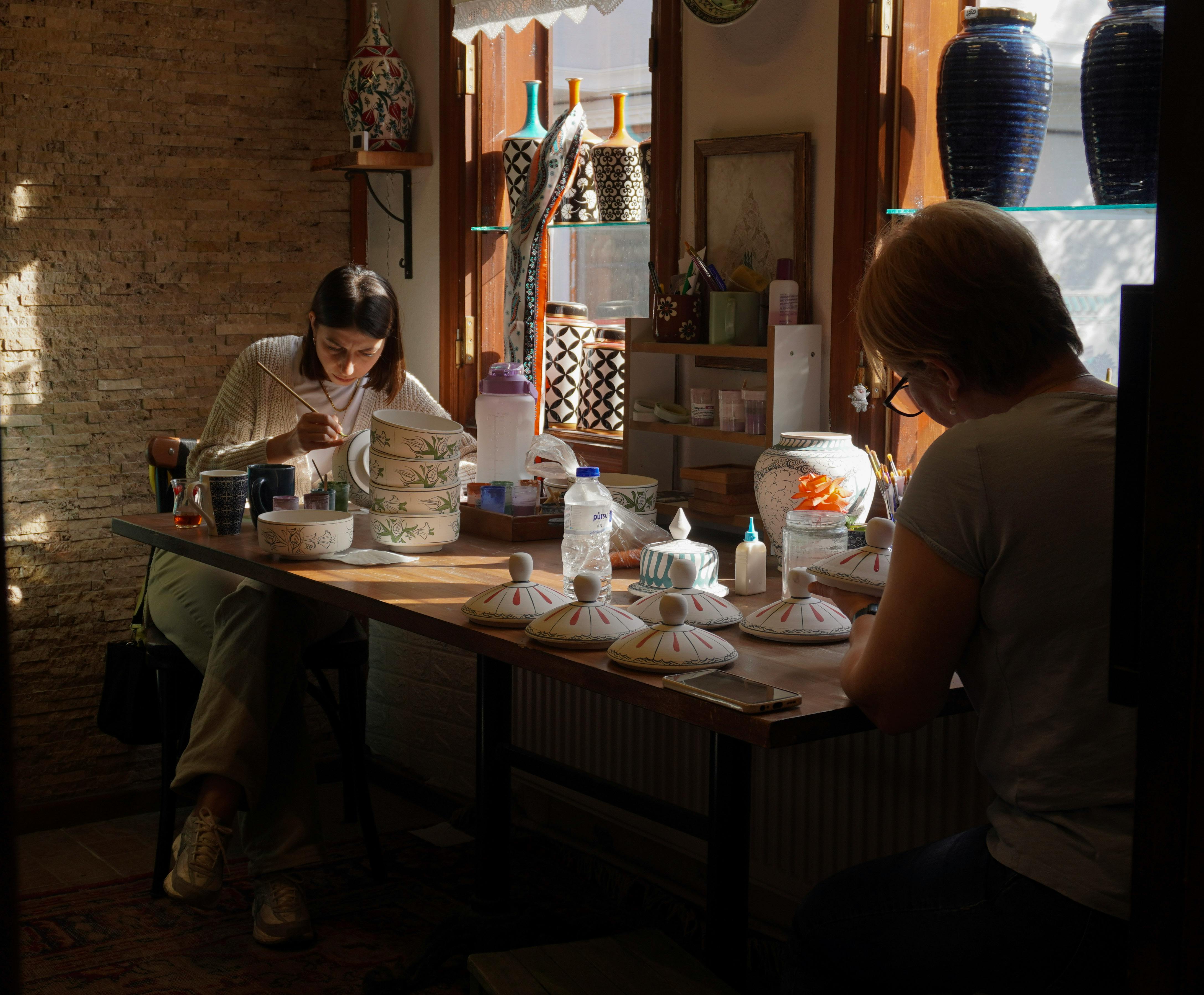 Artists hand-paint intricate designs on ceramics in a sunlit workshop in Bursa, Türkiye.