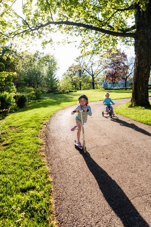 Free Two children joyfully riding scooters on a sunny day in a lush green park, creating a playful summer vibe. Stock Photo