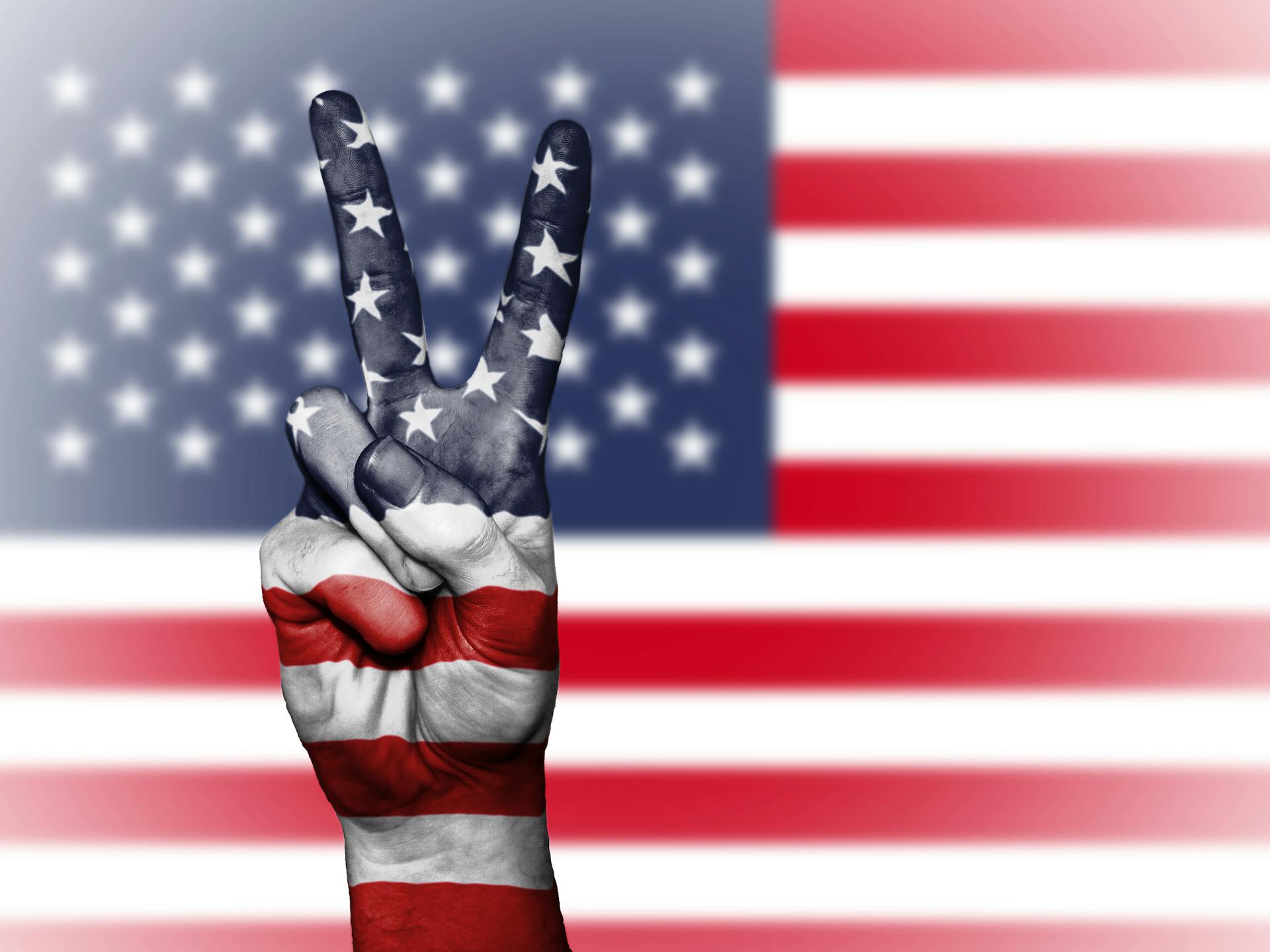 A peace sign hand painted with American flag colors against a blurred US flag backdrop symbolizing patriotism.