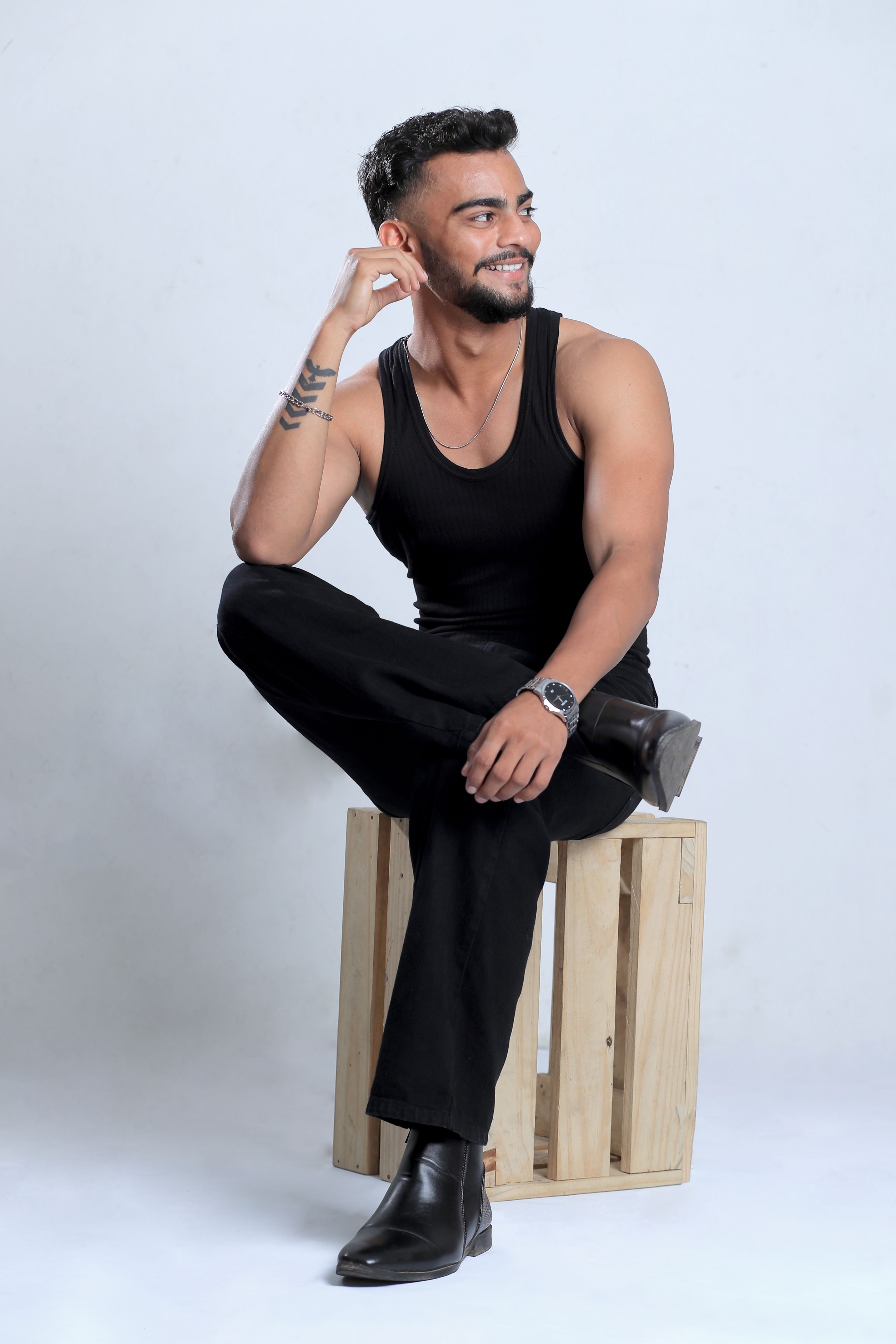 Stylish man in black tank top and pants posing on a wooden crate in a studio setting.