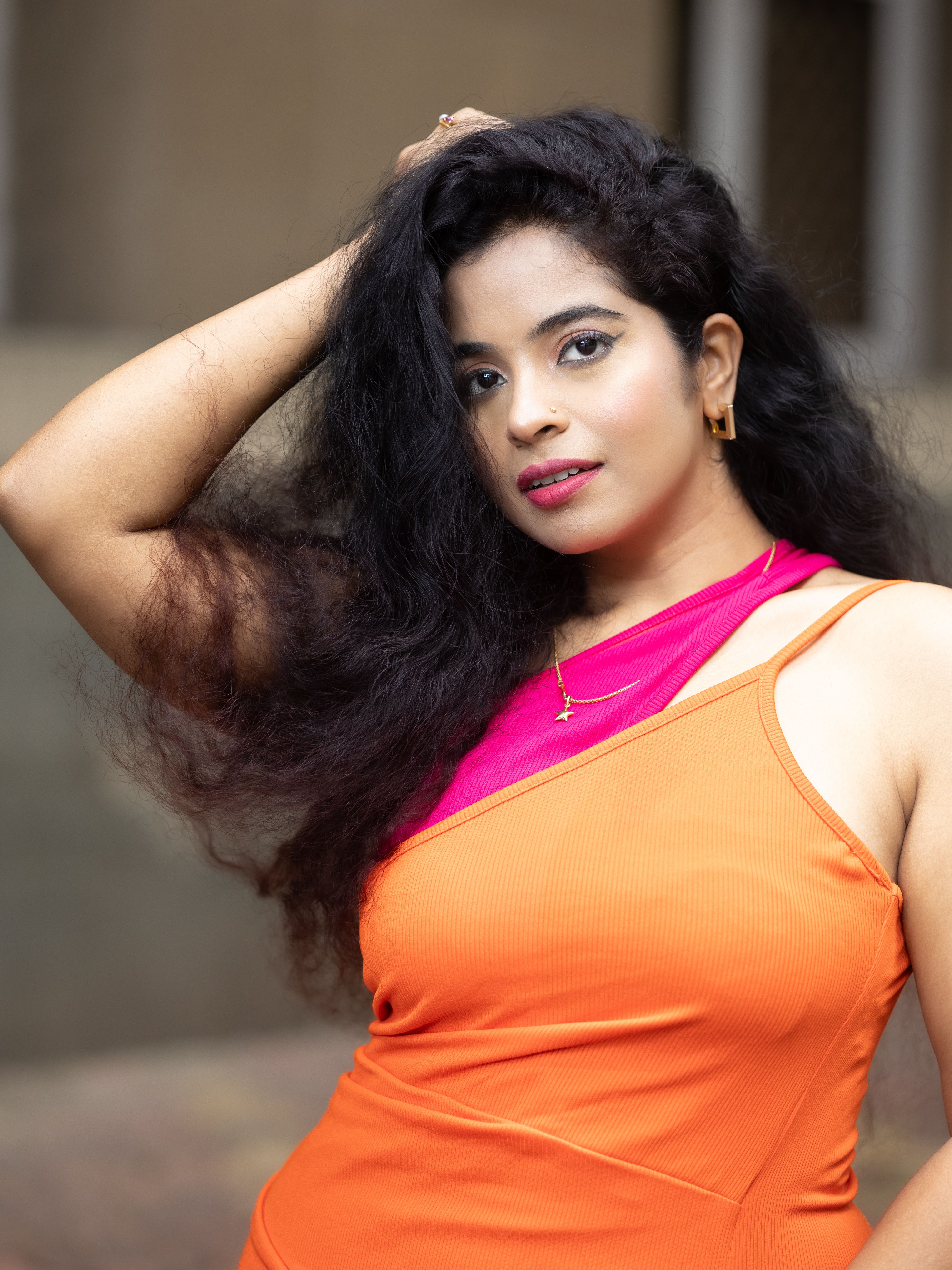 A fashionable woman in an orange dress poses confidently with her hand in her hair.
