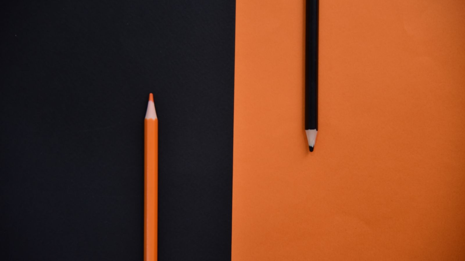 Two pencils, one orange and one black, lying on contrasting orange and black backgrounds