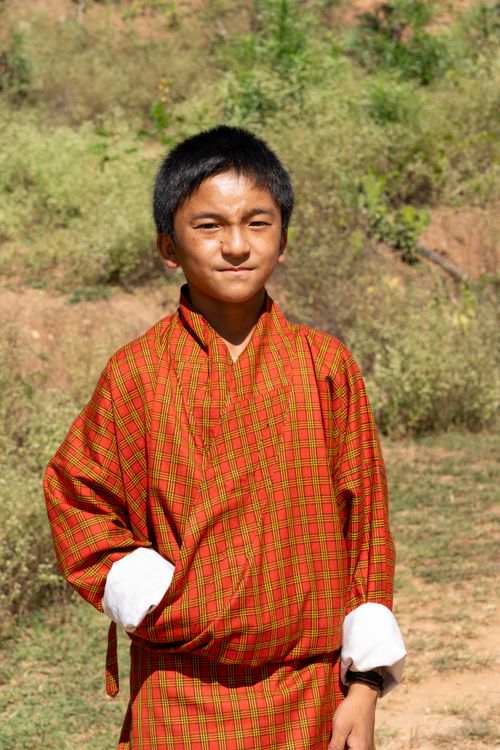 Free A young boy standing outdoors in traditional Bhutanese clothing, smiling confidently under the bright sun. Stock Photo