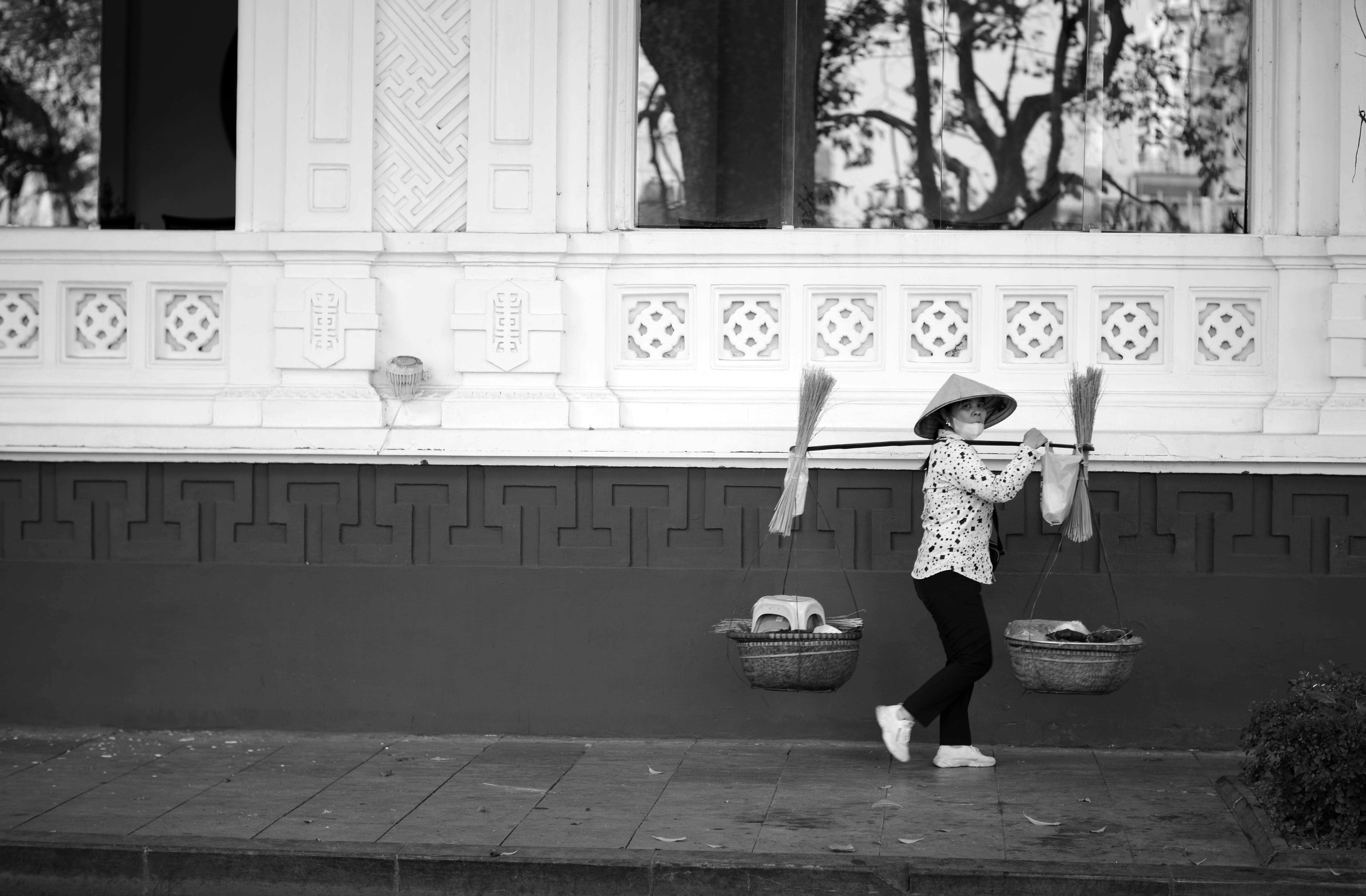 Street vendor carrying brooms in Hà Nội, with traditional conical hat and baskets.