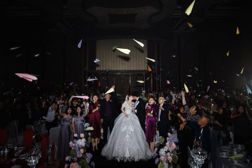 Free A joyful wedding celebration featuring a couple surrounded by guests tossing colorful paper planes indoors. Stock Photo