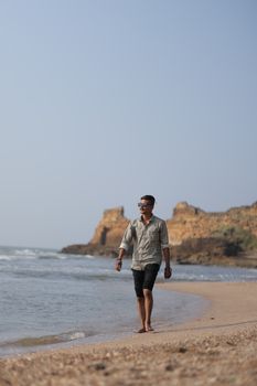 A man walks along a scenic beach with rocky cliffs under a clear sky.