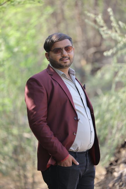 A fashionable man in sunglasses poses outdoors wearing a burgundy jacket and white shirt.