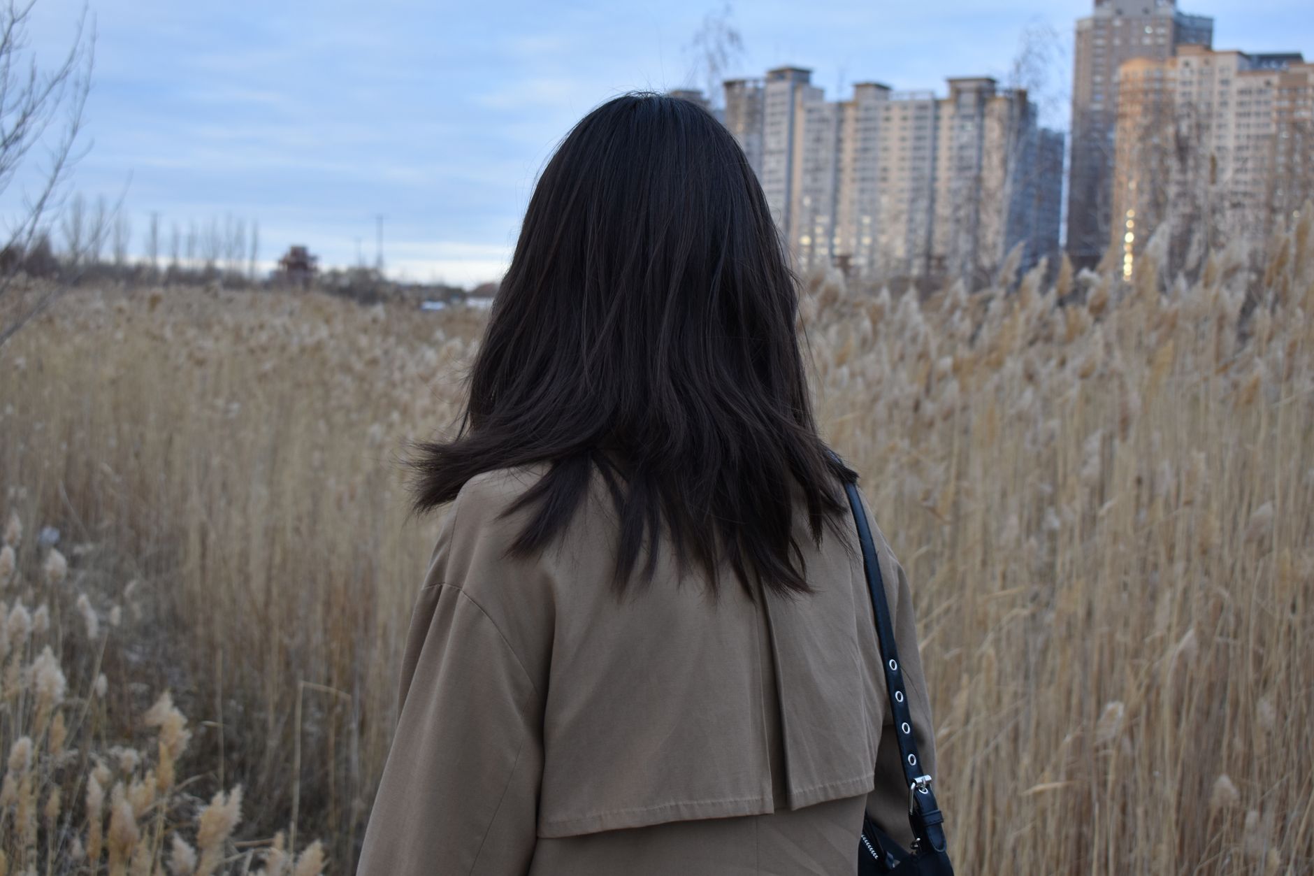 Woman standing in a field with city skyscrapers in Astana, Kazakhstan, wearing a beige coat.