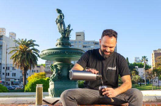 Man pours yerba mate in sunny Montevideo park with fountain backdrop.