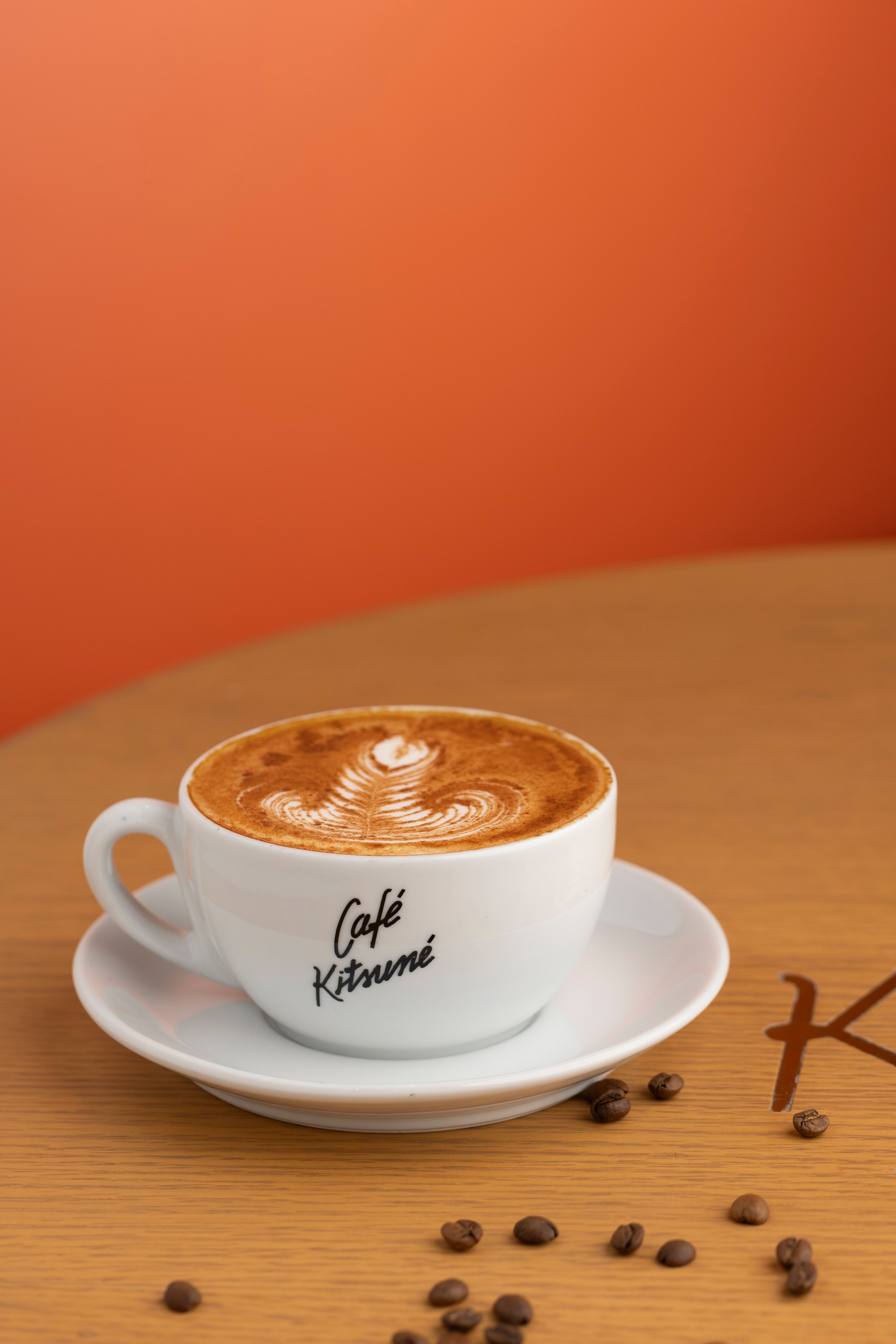 Elegant latte art in Café Kitsuné cup with coffee beans on a wooden table against an orange background.