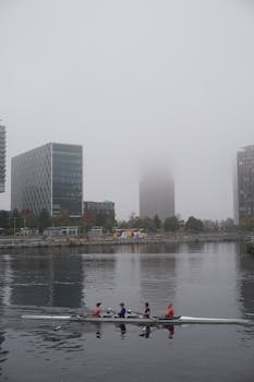 Rowers glide through misty waters of Salford Quays, with modern architecture in the background.