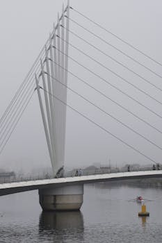 A foggy view of a modern bridge in Salford, England with rowers on the river beneath.