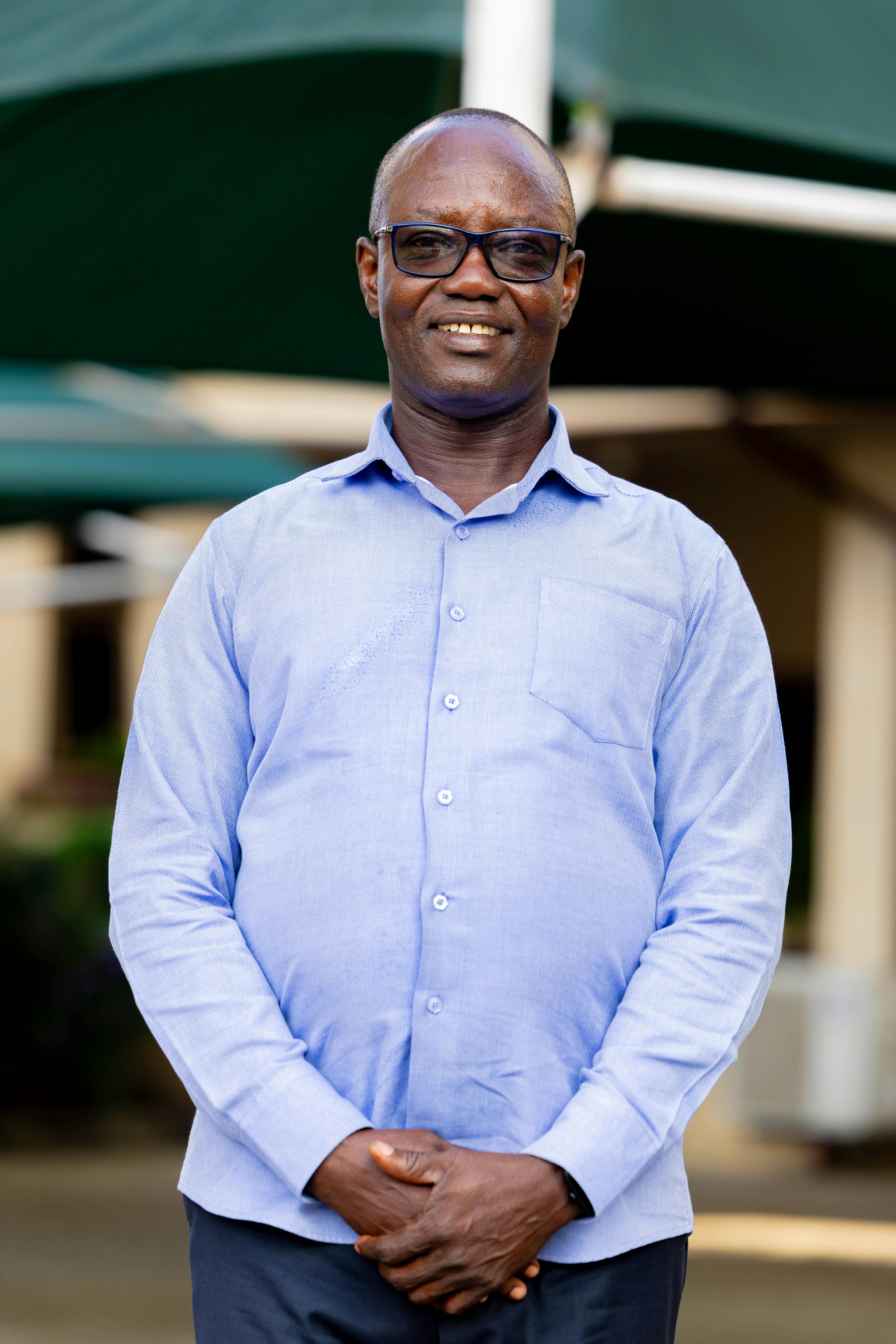 Smiling man wearing glasses and a blue shirt stands outdoors, conveying confidence and warmth.