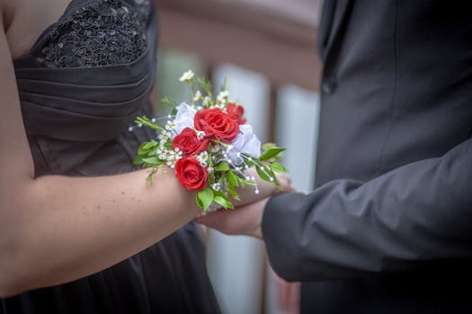 Una coppia in abito formale si tiene per mano, con un mazzo di rose rosse al polso della donna.