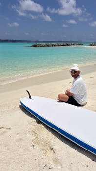 A man sitting next to a surfboard on the sunny shores of the Maldives.