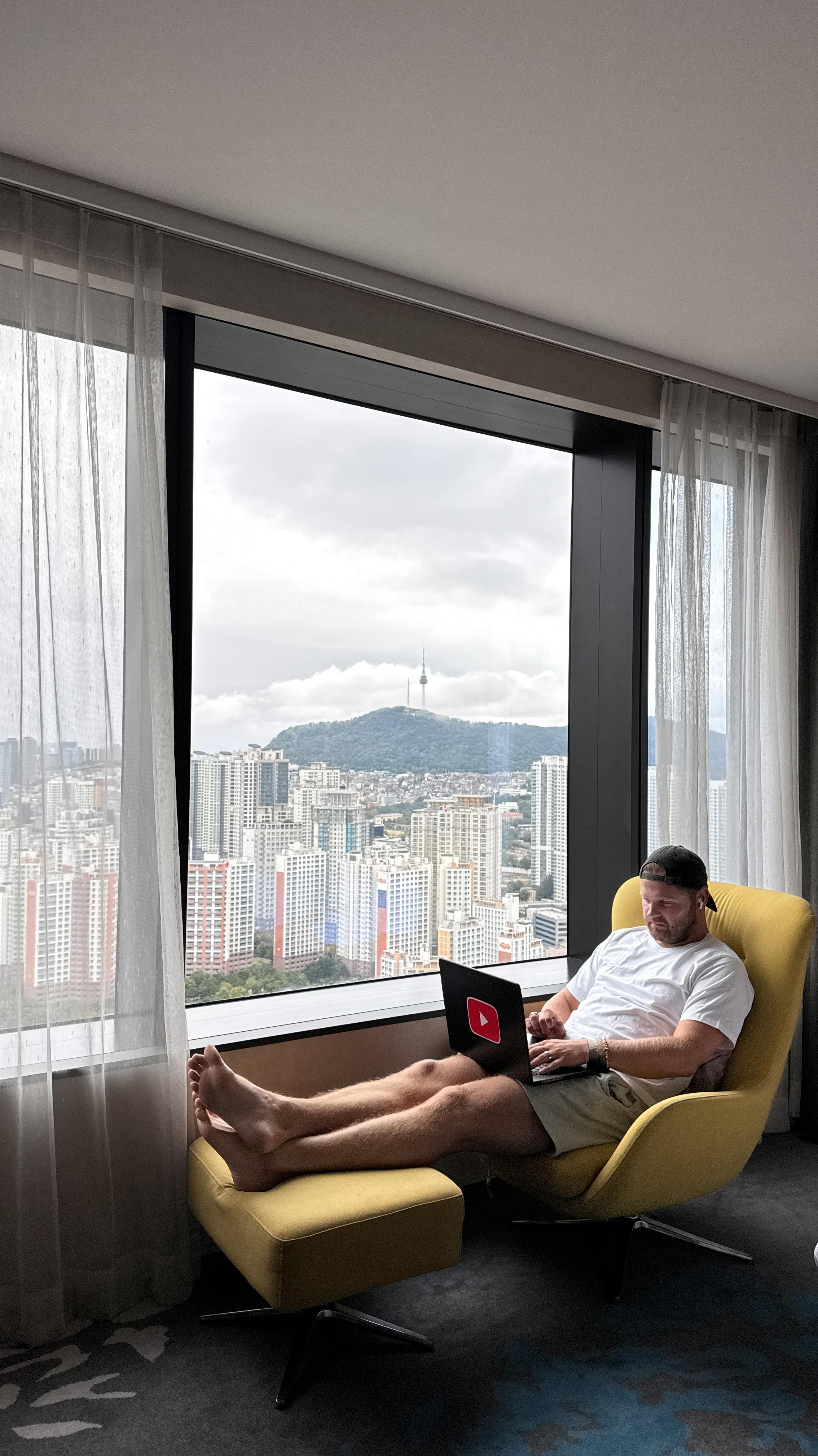 Man working on a laptop in a hotel room with a view of Seoul's skyline and Namsan Tower.
