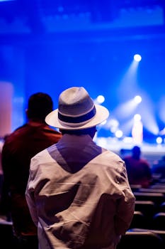 Person in white shirt and stylish hat watches a live performance in a vibrant, lit auditorium.