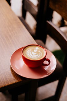 A red cup with latte art sits on a table in a cozy cafe setting, inviting warmth.