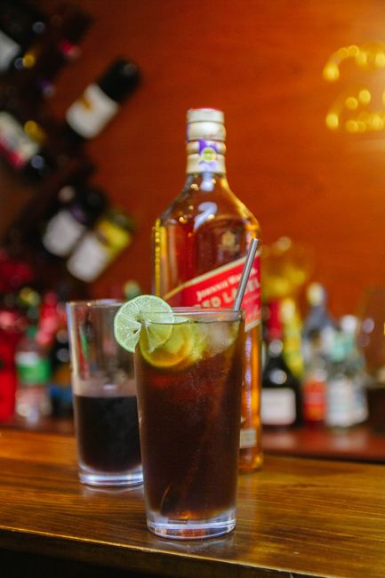 A tall glass of rum and coke filled with ice, dark cola, and a lime wedge on the rim, set on a wooden bar counter.