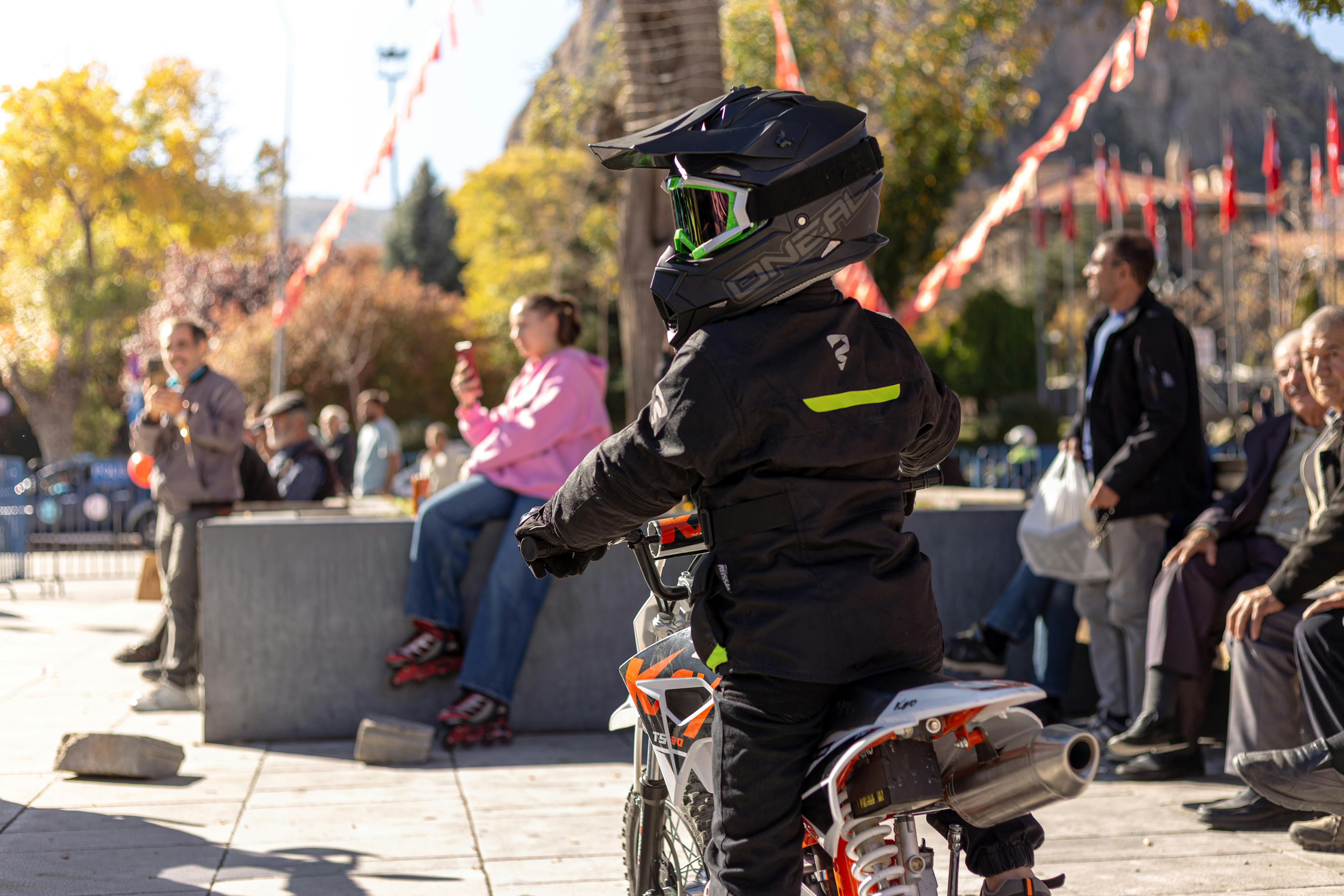 A child on a small motorcycle rides by spectators in a sunny park setting.