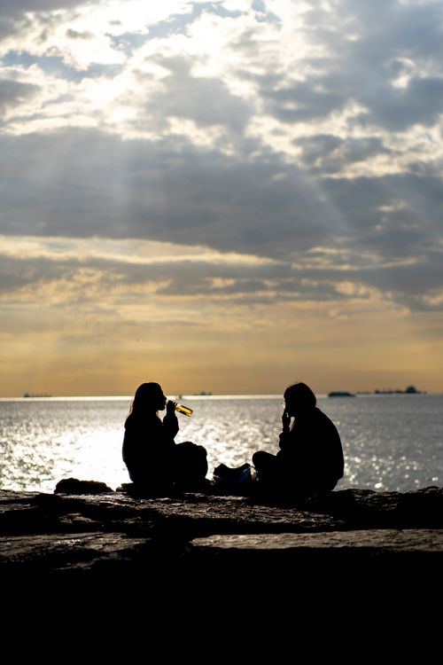 Free Two people sit by the ocean, silhouetted against a dramatic sunset sky. Stock Photo