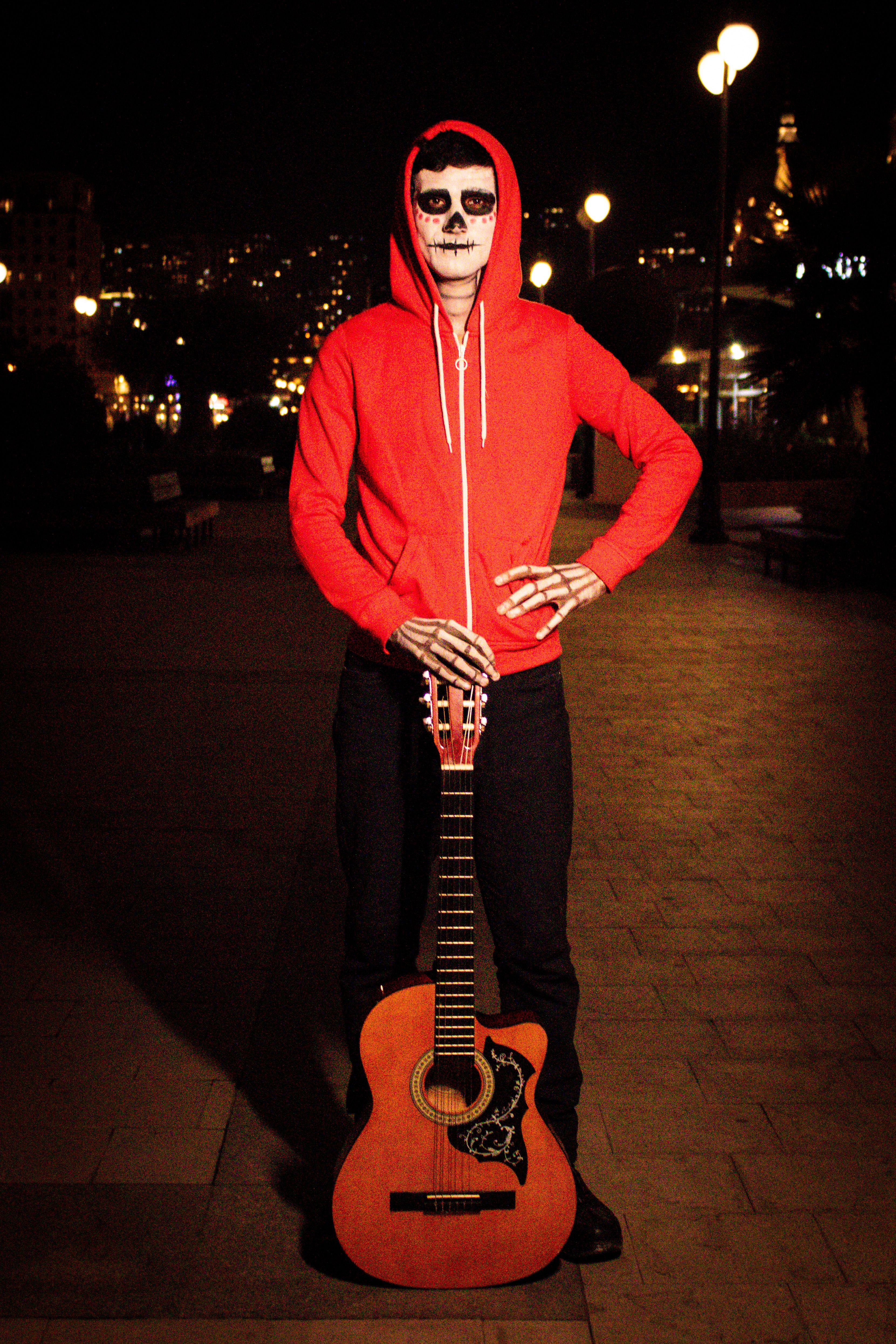 Person in red hoodie and skeleton makeup holding guitar at night outdoor setting.