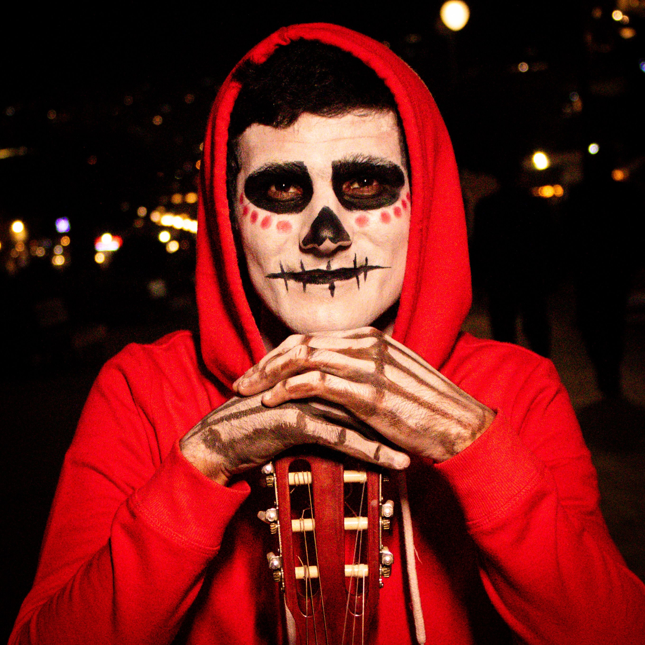 A person in a red hoodie with skull makeup holding a guitar, capturing spooky Halloween vibes at night.