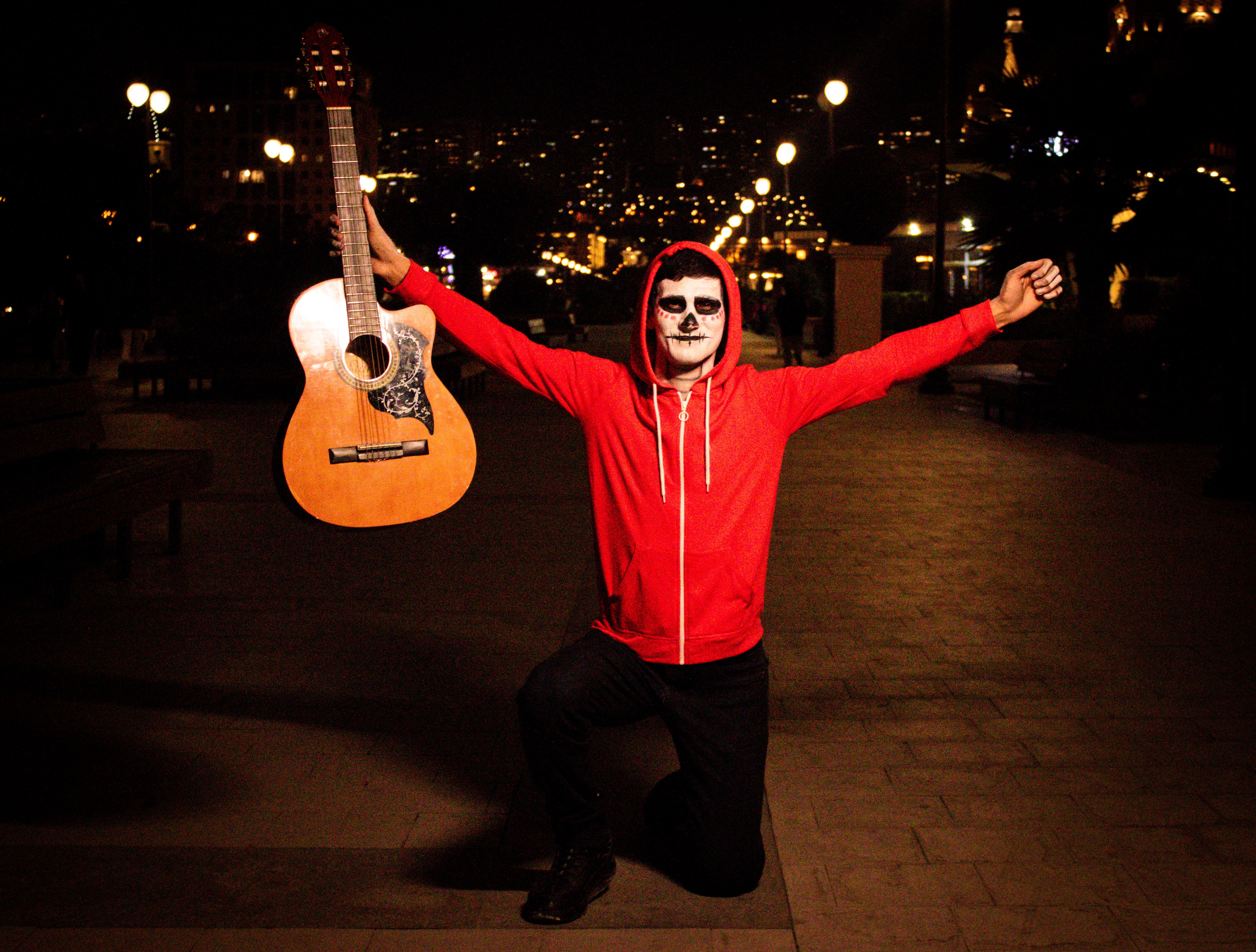 A man in skull makeup and red hoodie holds a guitar at night on a city street.