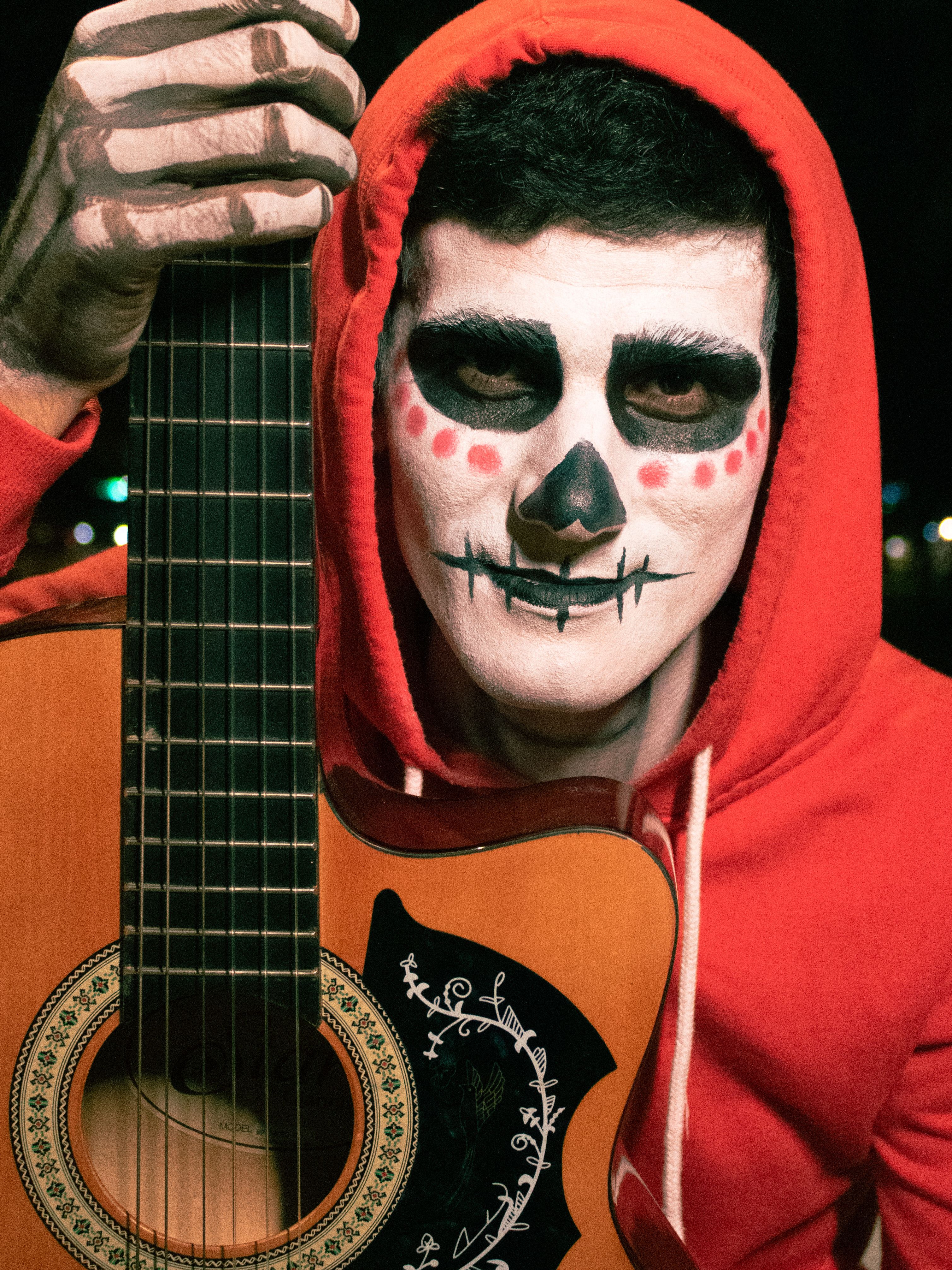 Guitarist in Dia de los Muertos makeup and red hoodie poses dramatically.
