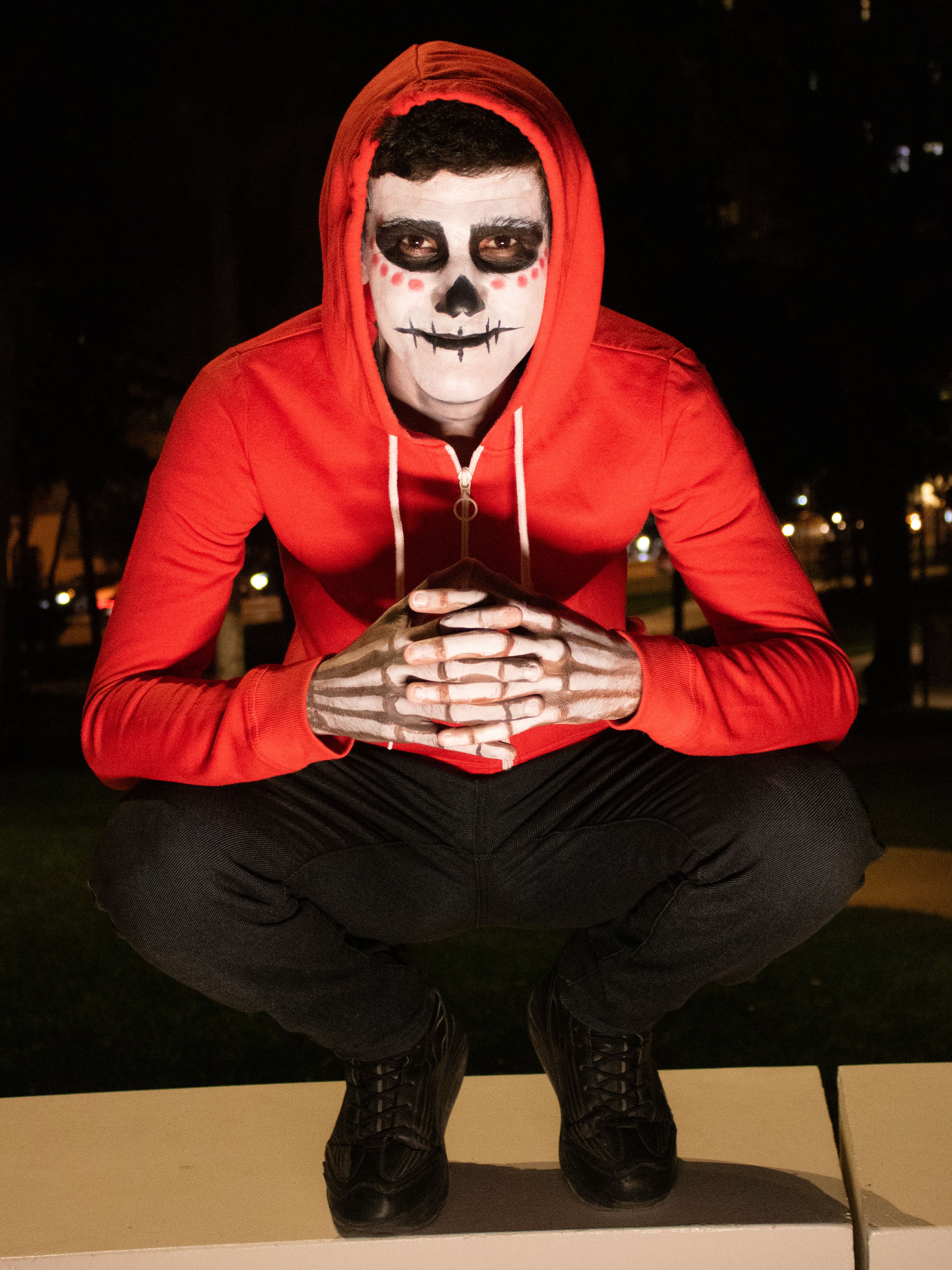 Man with skeleton face paint in a red hoodie crouching outdoors at night.