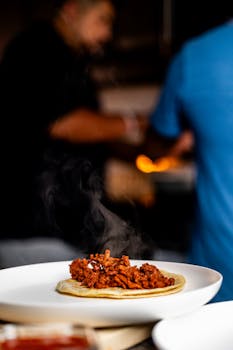 Steaming taco with meat filling on plate, chefs bustling in background, capturing culinary moment.